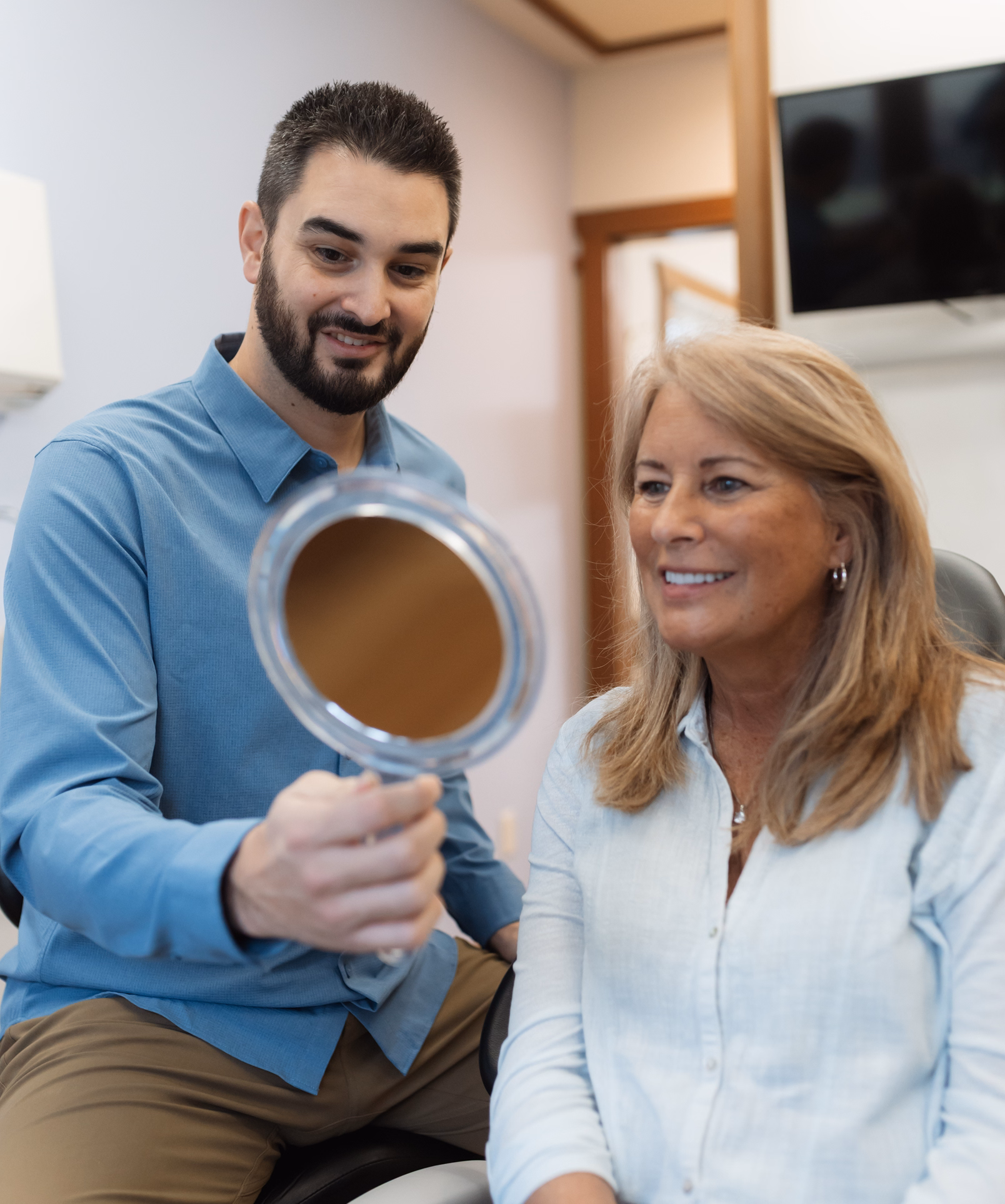 A man in a blue shirt holds a mirror for a smiling woman in a white blouse, seated in an office. The atmosphere appears friendly and relaxed.