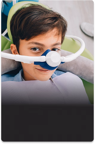 A child lies in a dental chair with a nitrous oxide mask over their nose, looking up.