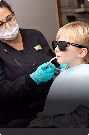 A child wearing sunglasses sits in a dental chair as a dental hygienist, in a black uniform and green gloves, uses a suction tool in a calm setting.
