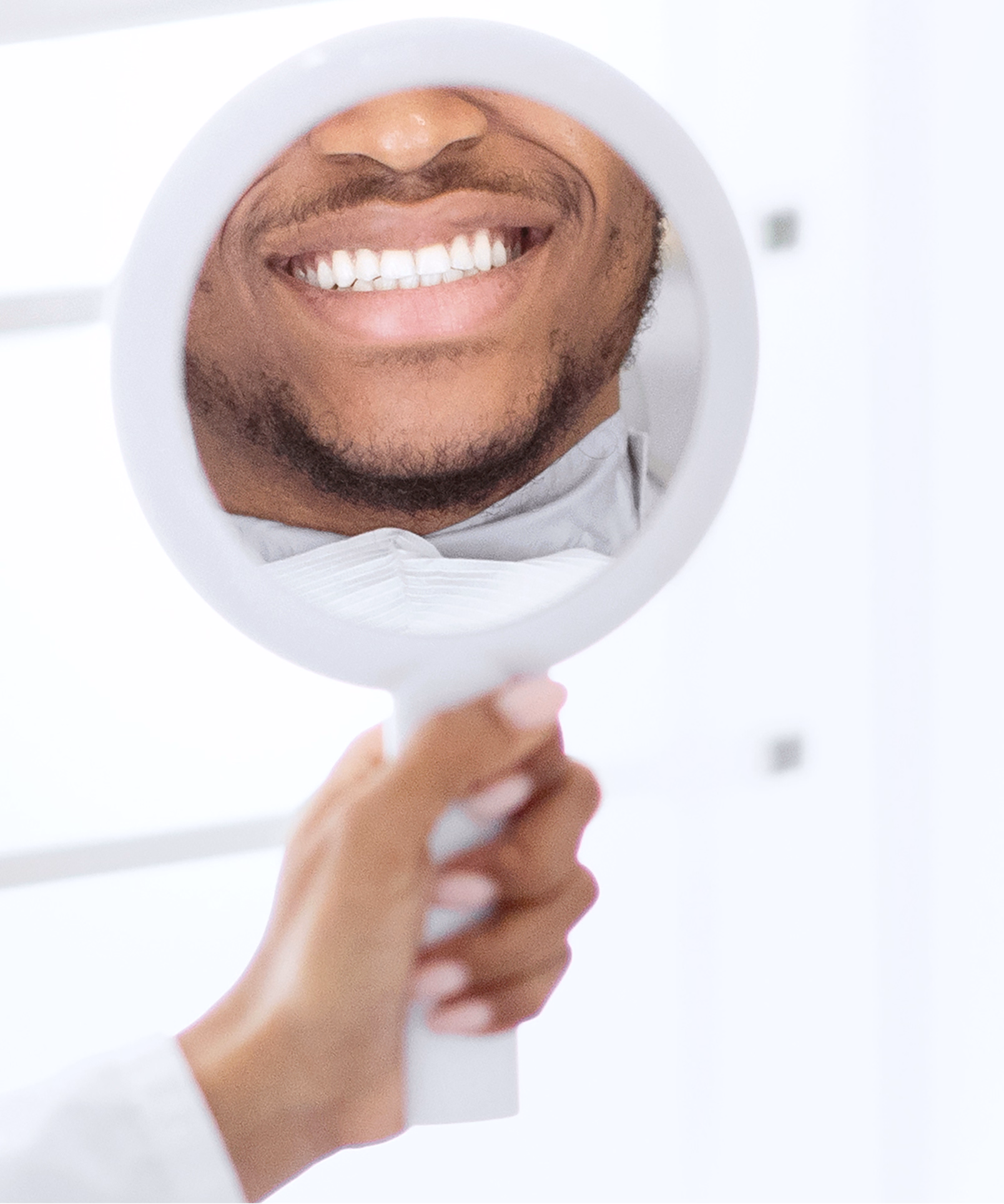 A person holds a round mirror, reflecting a bright, cheerful smile with perfectly white teeth. The scene is set in a well-lit, clinical environment.