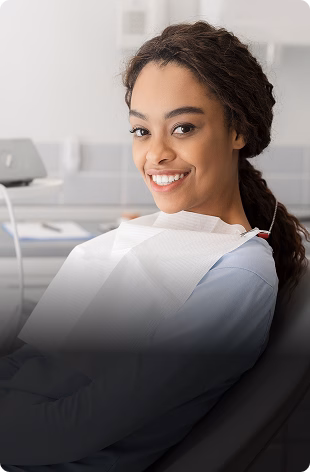 A smiling person with dark, braided hair sits in a dental chair wearing a dental bib. The setting is a clean, modern dental office, conveying a sense of calm.