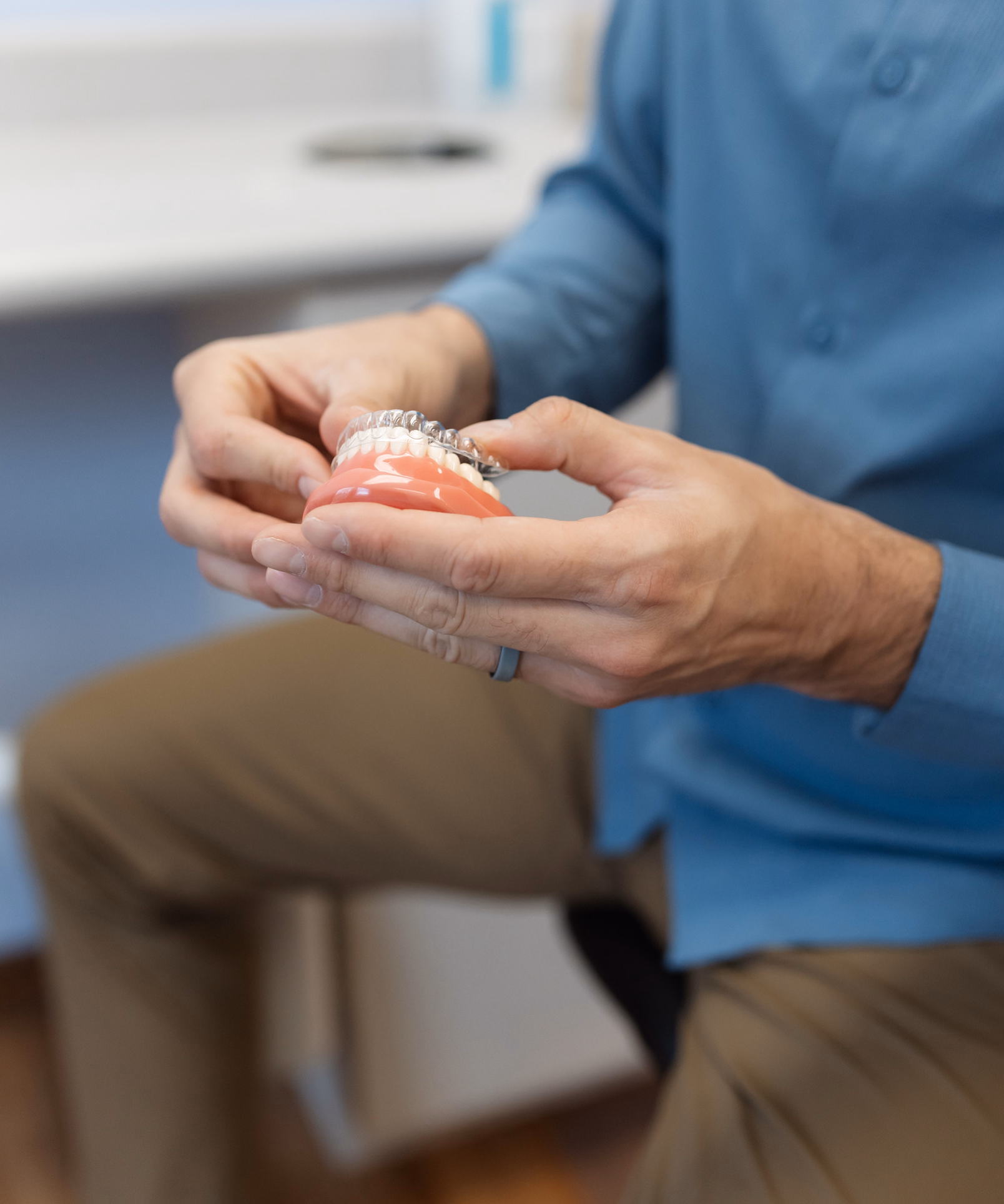 A person in a blue shirt holds a dental model showing teeth with braces, illustrating orthodontic care. The setting is a clean, professional office.