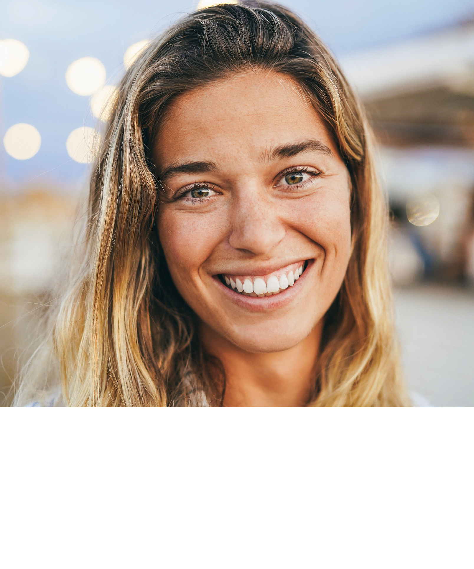 Smiling woman with long, wavy blonde hair and sun-kissed skin is close-up in soft focus. Bokeh lights and a tranquil evening backdrop enhance the warm, joyful tone.