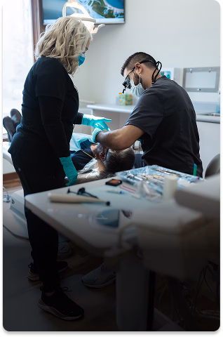 A dentist and assistant, wearing masks and gloves, work on a patient in a bright dental office. Dental tools are visible in the foreground.