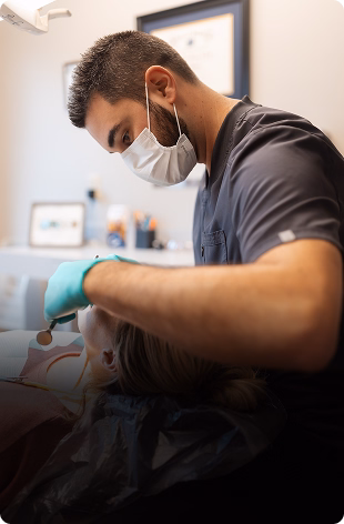A dentist wearing a mask and gloves performs a dental procedure on a reclined patient in a clinic. The setting is calm and professional.