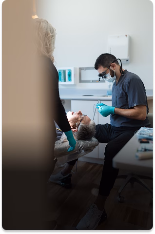 Dentist wearing gloves and mask examines a patient in a modern clinic. An assistant stands nearby. The atmosphere is professional and calm.