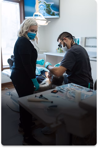 A dental office with two masked professionals treating a patient. The room is bright, with dental tools visible on a tray, conveying a clinical, focused atmosphere.