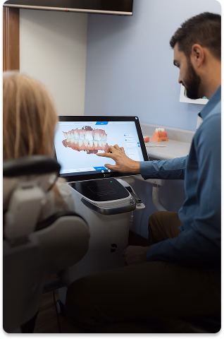 A dentist shows a patient a 3D dental model on a touchscreen in a calm clinic setting. The patient appears attentive, and the dentist explains details.