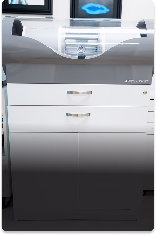 A sleek dental milling machine with a transparent cover sits atop a white cabinet. The room is clinical and modern, reflecting a professional setting.