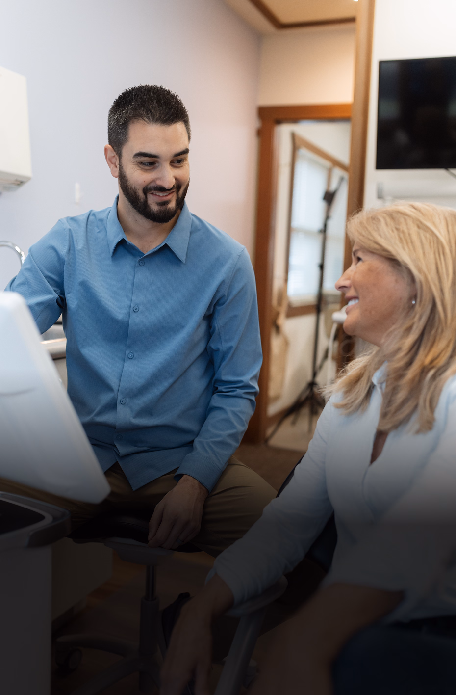 A man in a blue shirt sits beside a smiling woman in a dental office. They are engaged in a friendly conversation, creating a warm, welcoming atmosphere.