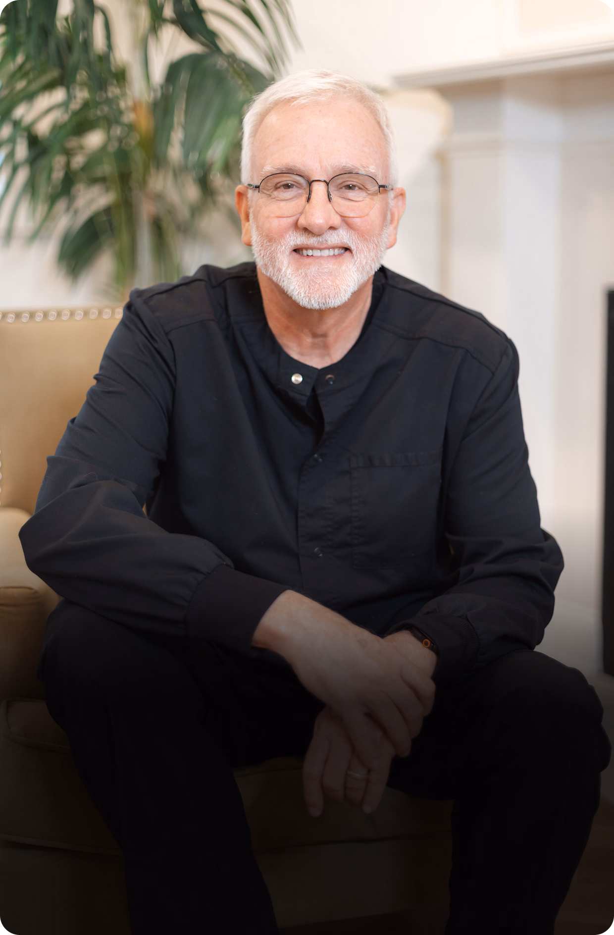 Elderly man with white hair and beard, wearing glasses and a dark shirt, smiles warmly while seated in a cozy room with a plant in the background.